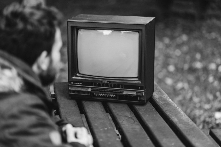 Black and white side view of anonymous male sitting at wooden table and looking at old fashioned television in nature on blurred background
